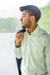 Fototapeta premium Dressing in a long sleeves shirt, a ivy cap, taking off his jacket, a young handsome black guy is standing by a lake in a raining, foggy day, smilingly looking at you..