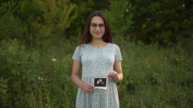 A young pregnant woman holds an ultrasound picture of a baby in her hands. Girl in glasses and a dress in nature.