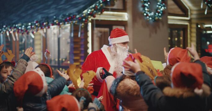 4K Santa Claus gives wrapped gift boxes to cute little children . Two Santa clauses helping each other to give gifts to queue of children . Many kids standing and waiting in line queue for a gift .