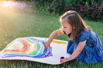 Obraz na płótnie Canvas Little girl 2-4 years old paints rainbow and sun on large sheet of paper, sitting on green lawn in sunlight