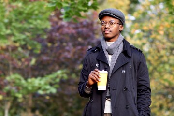 Portrait of handsome serious black guy, young African Afro American ethnic man in hat, scarf and coat in glasses is walking at golden autumn park at beautiful fall with a cup of tea or coffee to go