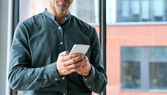 Close up of businessman holding using cell mobile smartphone in hands reading in office near panoramic window. Technology applications for business development growth and solutions concept.