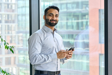 Smiling indian ceo businessman holding using cell phone mobile apps standing in office near...