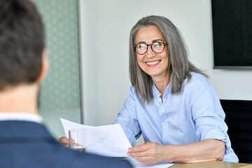 Smiling happy mature ceo businesswoman wearing glasses looking listening to executive manager discussing corporation financial plan at office table holding documents. Job interview.