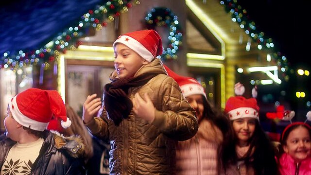 Happy children are jumping , laughing and looking camera into front of the beautiful house decorated with lights and garlands. Portrait of many kids with Santa hat and costumes at Christmas night .