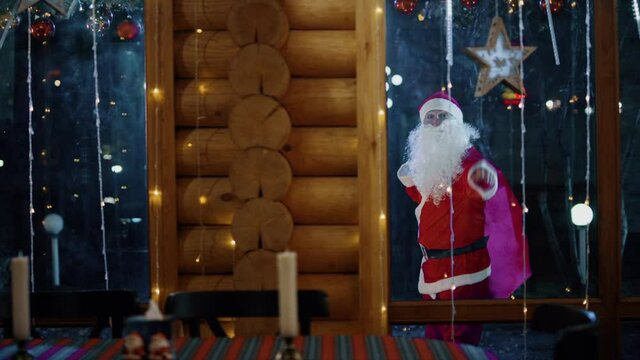 Happy Santa Claus walking and showing hand wave near the wooden home with large bag . Funny santa clause walking on the street with large sack at night . Outside the window . Slow Motion . Say hi .