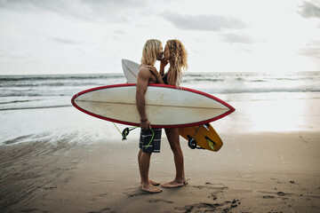 Handsome man and his girlfriend are kissing against background of sea and holding surfboards