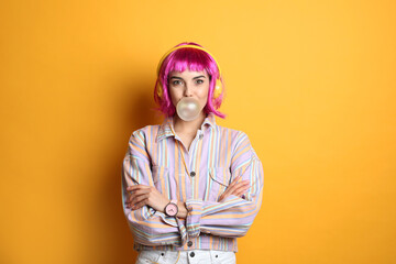 Fashionable young woman in colorful wig with headphones blowing bubblegum on yellow background