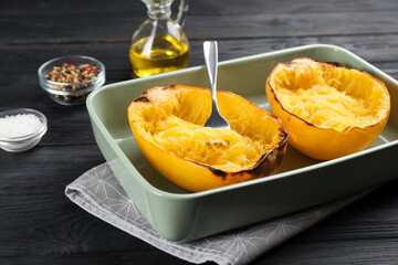 Halves of cooked spaghetti squash and fork in baking  dish on black wooden table, closeup