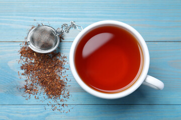 Ceramic cup of aromatic rooibos tea, infuser and scattered dry leaves on light blue wooden table,...