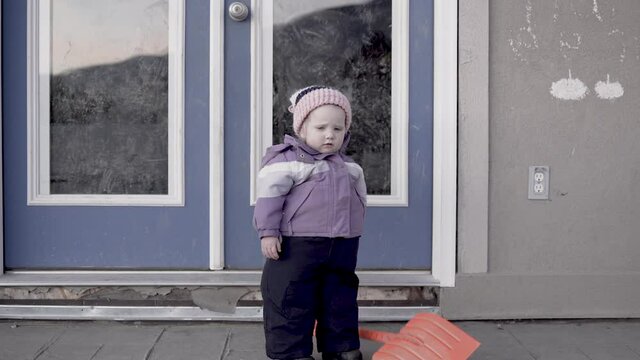 Small child waits outside with a snow shovel in a snowless winter