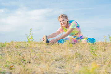 A young beautiful girl in a bright tracksuit sits on green grass against a blue sky and performs physical exercises to stretch the muscles. Outdoor sports.