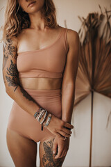 Tanned blonde woman in beige retro style swimsuit poses near palm leaf. Photo of cheerful girl with tattoos on hand and handmade bracelets