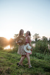 Two young beautiful smiling women in summer dresses and with straw hats posing in front of lake on warm summer day at sunset. Best girlfriends outdoors. Positive models having fun and hugging in park