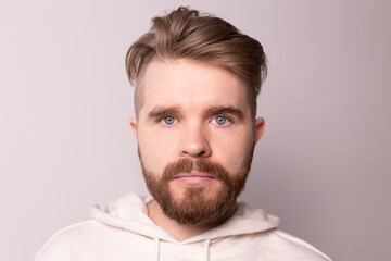 Portrait of young male with beard and mustache and trendy hairdo. Wears casual hoodie, has serious expression in studio against white background