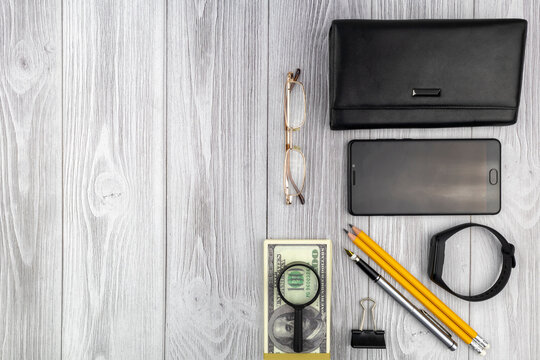 Office financial flat lay still life with 100 US Dollar bills, magnifying glass, purse, smartphone and stationery over a grey wood background with copyspace