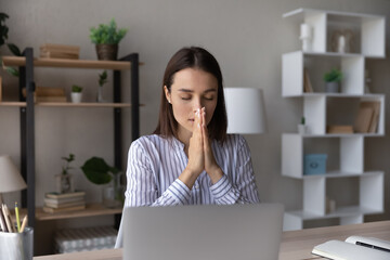 Superstitious millennial Caucasian woman work on computer hold hands in prayer ask beg for good...