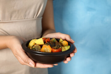 Woman holding earthenware with delicious roasted potatoes and meat on light blue background, closeup