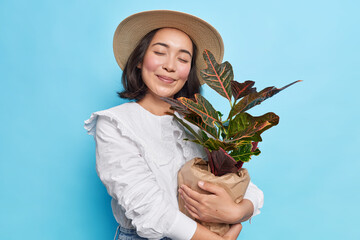Pleased young Asian woman closes eyes embraces pot of houseplant happy to buy new flower for her home garden wears hat white blouse isolated over blue background. People and lifestyle concept - Powered by Adobe
