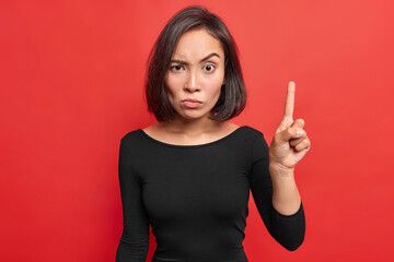 Serious strict Asian woman with dark hair raises index finger looks with angry expression warns you...