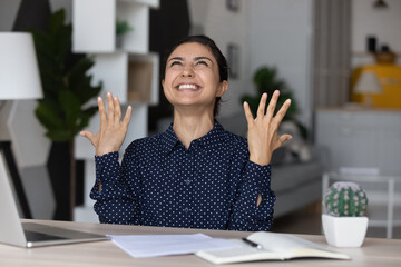 Overjoyed young Indian woman sit at desk at home office work on laptop feel euphoric with good...
