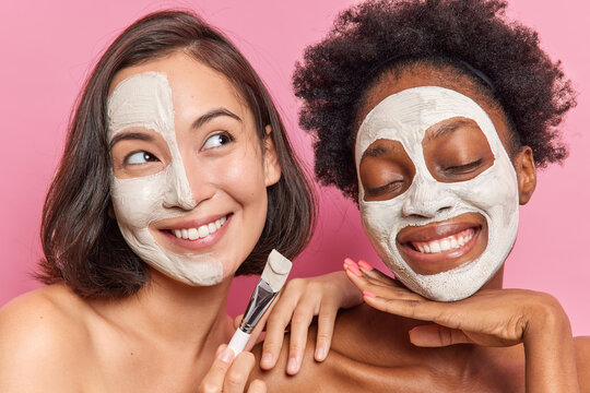 Photo of smiling good looking multiethnic women apply nourishing mask on facew with cosmetic brush stand shirtless dream about something pleasant isolated over pink background. Beauty concept