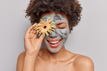 Pleased curly haired woman applies clay mask on face for skin rejuvenation holds flower over eye...