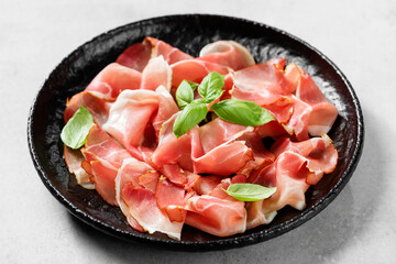 Tasty prosciutto slices with basil leaves on light gray stone background. 