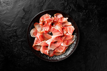 Slices of prosciutto di parma or jamon serrano  on a black plate on a black stone  background. Top view .