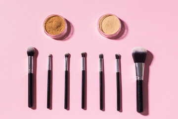 Various make-up brushes and face mineral powder on pink background with copy space, top view. Cosmetics and beauty concept.