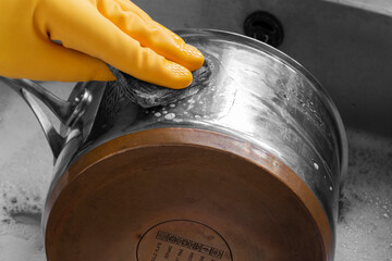 Person, wearing rubber gloves, cleaning a stainless steel and copper saucepan with a wire pad.