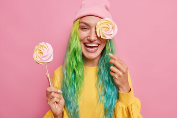 Positive teenage girl holds two round candies on sticks covers eye with delicious caramel lollipop has colorful hair wears yellow jumper and hat isolated over pink background being in good mood