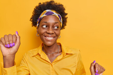 Smiling carefree dark skinned woman with natural curly hair has upbeat mood dances and keeps arms raised wears headband shirt poses against vivid yellow background. People fun lifestyle concept