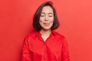 Pretty brunette young Asian woman closes eyes licks lipsfrom temptation to taste something delicious shows tongue imagines eating delicious food wears shirt isolated over vivid red background - obrazy, fototapety, plakaty