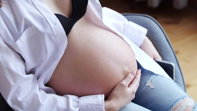Young pregnant woman stroking big belly on armchair. 