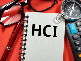 Text HCI (Human Computer Interface) writing on notebook with compass,pen,calculator and glasses on red background.