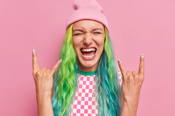 Joyful millennial woman makes rock n roll gesture shows she is cool feels very happy has long dyed...