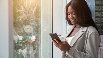 Positive African-American business lady in stylish jacket drinks coffee and texts on mobile phone by large window having break in office, sunlight