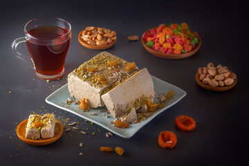 Sunflower halva sprinkled with crushed nuts and raisins on a blue plate with pistachios, peanuts,dried apricots, candied fruit syrup and a glass of tea on a dark background. Eastern sweets