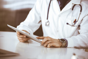 Unknown male doctor sitting and working with tablet computer in clinic at his working place, close-up. Young physician at work. Perfect medical service, medicine concept