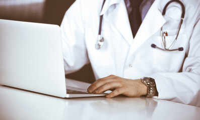 Unknown male doctor sitting and working with laptop in clinic at his working place, close-up. Young physician at work. Perfect medical service, medicine concept