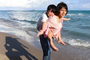 Mother piggybacking her kid on beach, playing together on summer holiday vacation, joyful parent with child boy spending time on tropical sea beach. Happy family on weekend