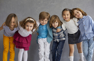 Little friends having fun together. Group portrait of happy young children. Team of positive kids in casual wear standing against grey studio wall, hugging, huddling, smiling and looking at camera