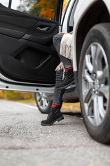 Fototapeta na wymiar Stylish unrecognizable girl in grey jeans, beige scarf and black boots sitting in car doorway. One foot staying on asphalt. Selective focus on leg. Autumn season, colorful background. Roadtrip concept