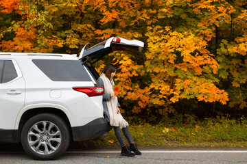 Attractive brunette woman in grey coat, black boots and beige scarf sitting in car trunk. In hand she hold multicolored maple leaves. Picturesque autumn season. Travelling by car. Beauty in nature. 