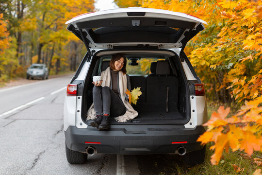 Happy caucasian girl sitting in SUV trunk and hold cup with tea. Stopping in a beautiful place while car trip. Girl enjoying autumn vacation. Golden foliage in blurred background. Traveler lifestyle.