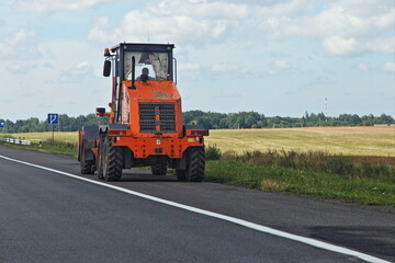 Big orange wheeled tractor bulldozer drive on rural asphalted road side on field and blue sky background at summmer day, East Russian farm landscape