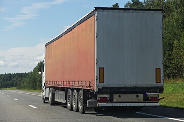 One European brown semi trucks back side view on free suburban highway road at Sunny summer day on green forest background, cargo transportation logistics traffic landscape