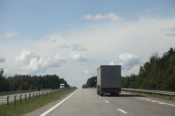 European heavy truck move on the countryside highway road at Sunny summer day on green forest, road fence and blue sky with clouds background, back side view, transportation logistics