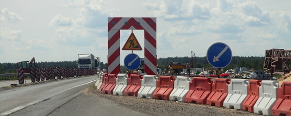 Detour of the road repair site, traffic direction arrow sign and road works sign on white red fence on right lane of the suburban highway at summer day